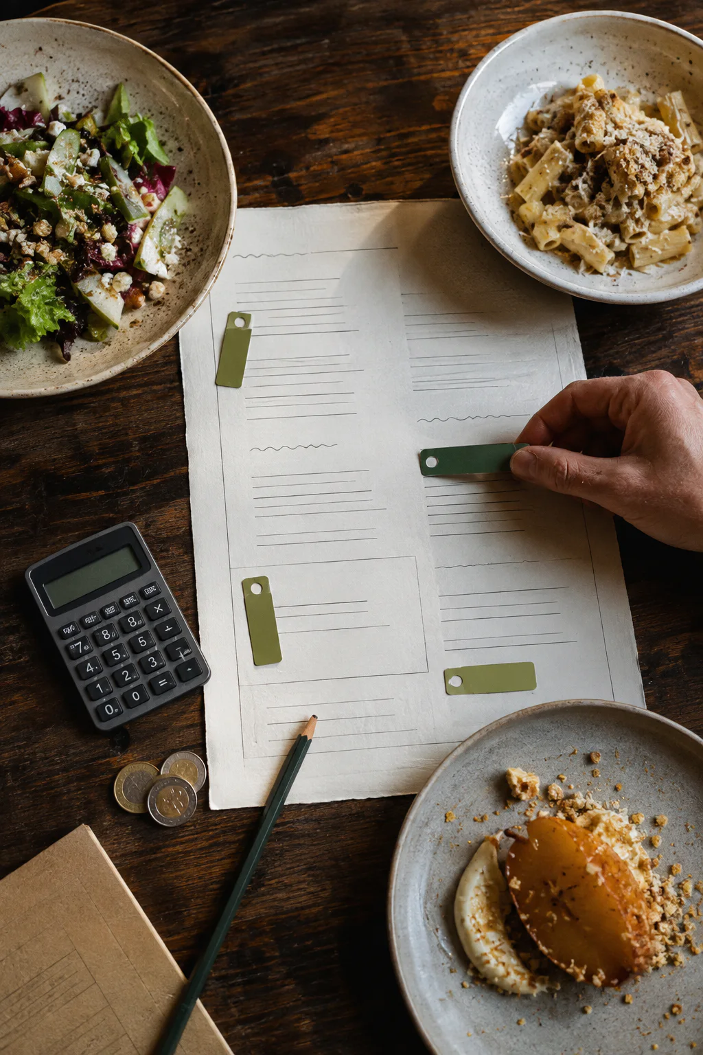 A hand arranges markers on a menu draft surrounded by dishes, a calculator, and coins.