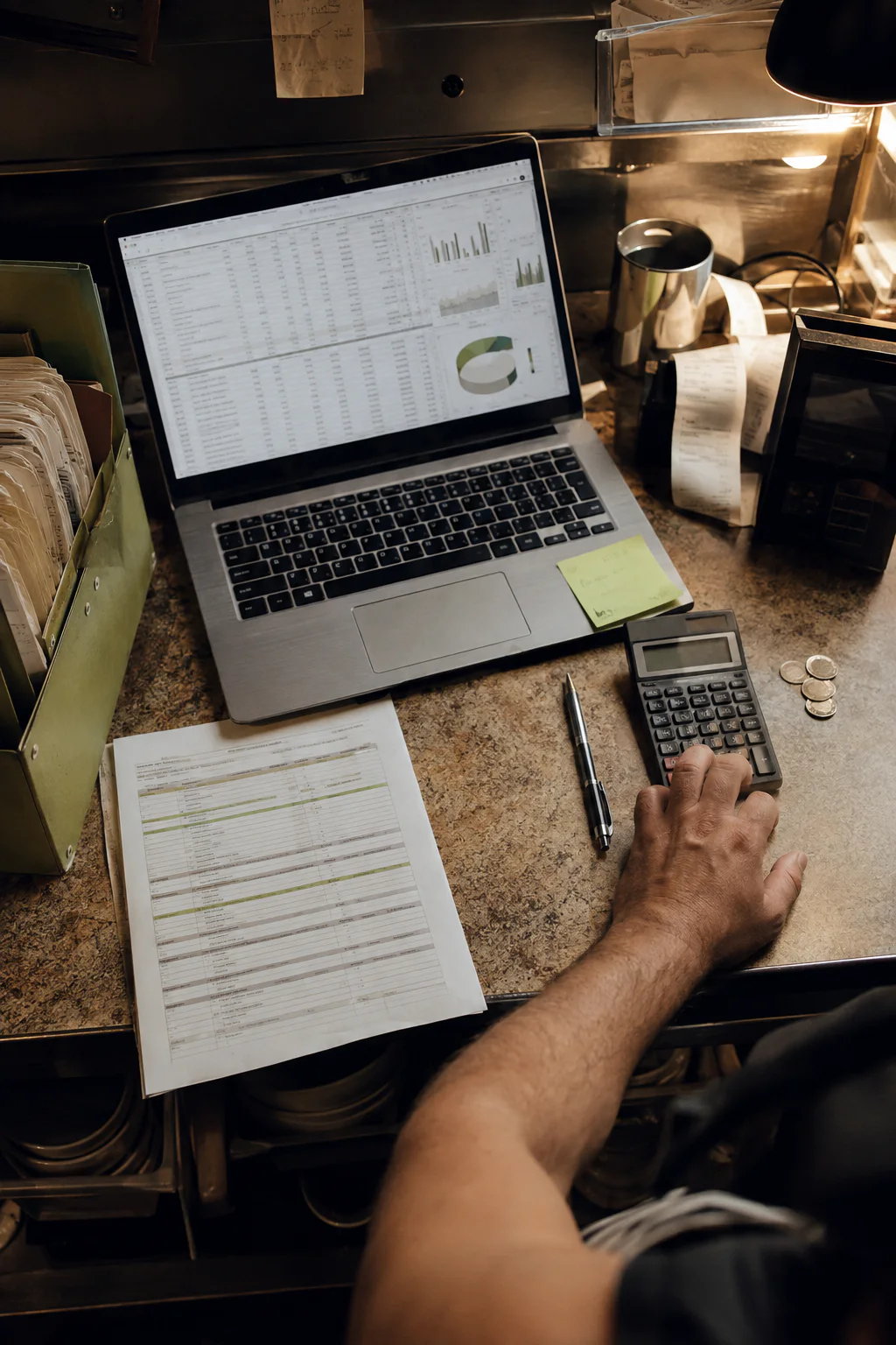 A hand calculates results beside a computer with a restaurant operations spreadsheet, receipts, and records.